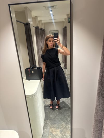 The image captures a woman taking a selfie in a dressing room. She's dressed in a black top and a flowing black skirt. She holds her smartphone in her left hand, positioning it to capture her reflection in the mirror. Her right hand is adorned with multiple gold bracelets. She has long, light brown hair and is looking directly at her phone screen. Beside her is a black Chanel handbag with gold hardware and a red and green ribbon detail. The dressing room has a bench seat, grey tiled flooring, and vertical lighting fixtures. The mirror reflects a corridor with dressing rooms to the right. While the exact location in Leiria, Portugal cannot be determined from this image alone, the presence of a dressing room suggests it's within a clothing store or boutique.