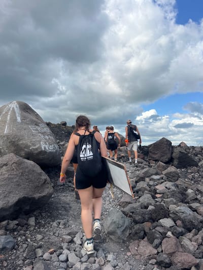 The image depicts a group of people on a rocky, volcanic terrain. The subjects appear to be hikers or adventurers, dressed in casual attire, including t-shirts, shorts, and athletic shoes. Some are wearing backpacks. The scene is set on a mountain or volcanic slope, characterized by a rugged landscape of dark rocks and sparse vegetation. The sky above is partially cloudy, with a mix of blue sky and gray clouds, suggesting a dynamic weather condition.

The activity taking place seems to be a hike or exploration of the volcanic area. One person in the foreground is being photographed, while others are walking ahead. There's a whiteboard or similar flat object being carried by someone in the middle ground. The overall mood seems casual and exploratory, with the subjects focused on their surroundings and the activity.

Notable details include the time of day, which appears to be daytime based on the lighting and sky. The location is suggested to be Rota, Nicaragua, which is known for its volcanic landscapes. There is some visible text on the backpacks of the individuals, featuring logos and what appears to be a website address or name. Additionally, a large boulder in the foreground has white markings on it, possibly graffiti or trail markers. The terrain's rough texture and the presence of numerous rocks are prominent features of the setting.