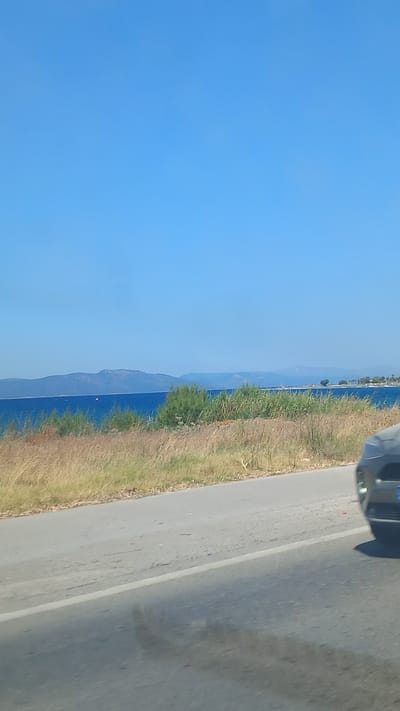The image depicts a roadside view with a body of water and distant landmasses. A gray car is partially visible in the lower right corner, driving on an asphalt road. The road has a white line marking. To the left of the road is dry, yellowish vegetation and some green bushes. Beyond the vegetation, a body of blue water is visible, with mountains or hills on the horizon. The sky is clear and blue. The scene suggests a coastal area.