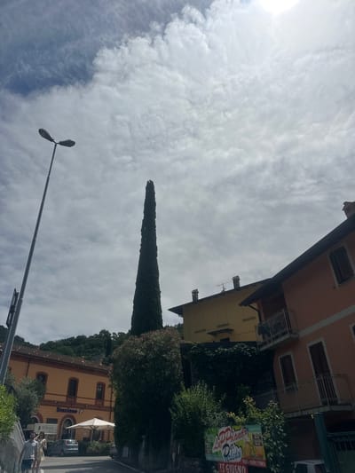 The image shows a street scene in Gittana, Italy. On the left, a tall lamppost stands against a cloudy sky. Behind it, a yellow building with arched windows displays the word "Stazione f," indicating it is a train station. A few people are walking on the sidewalk in front of the station. Further to the right, a very tall, slender cypress tree dominates the scene. Behind the tree, two multi-story buildings are visible: a yellow one with visible chimneys and a reddish-orange one with balconies and shutters. A sign with an advertisement is visible on the lower right side of the image, featuring images of beer and text. The overall setting appears to be a town, possibly with a Mediterranean feel, given the architecture and the lush greenery in the background.