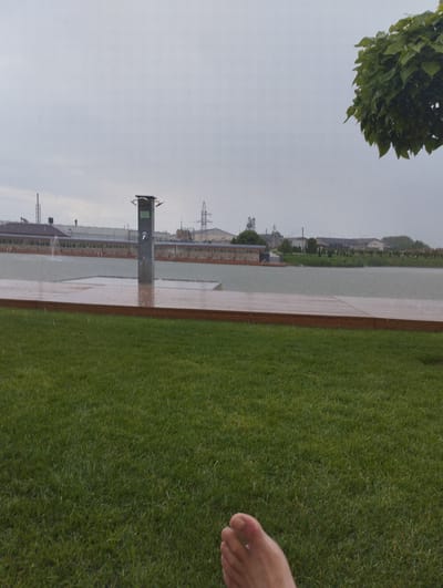 A person's foot is visible in the foreground, resting on a lush green lawn. The background depicts a slightly overcast day, with light rain falling, creating ripples on the surface of a tranquil body of water. A wooden deck, also wet from the rain, extends towards the water. In the middle ground, a modern outdoor shower stands next to the water. Further beyond, a long, low building with a dark roof is visible, with various industrial structures and power lines on the horizon. A large, leafy tree branch overhangs from the upper right corner of the frame, its leaves also speckled with raindrops. The overall scene suggests an outdoor recreational area, possibly a spa or a resort, during a rainy day. The weather is gloomy, but the presence of the lawn and the shower implies the potential for outdoor activities. No visible text is present in the image.