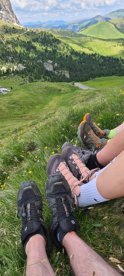 The image shows a close-up view of four people's legs and hiking boots resting on a grassy hillside. The background features a sweeping landscape of green rolling hills dotted with pine trees, a winding road, and distant mountains under a cloudy sky. The location is identified as Sëlva - Wolkenstein - Selva di Val Gardena, Italy, a region known for its mountainous terrain and hiking trails. The people are wearing various hiking boots, including black Salomon boots, pink and black boots, and green and orange boots. Some are wearing socks, one pair being white and another lime green. The grass is green and interspersed with small yellow wildflowers. The scene suggests a break during a hike, with individuals relaxing and enjoying the scenic view.