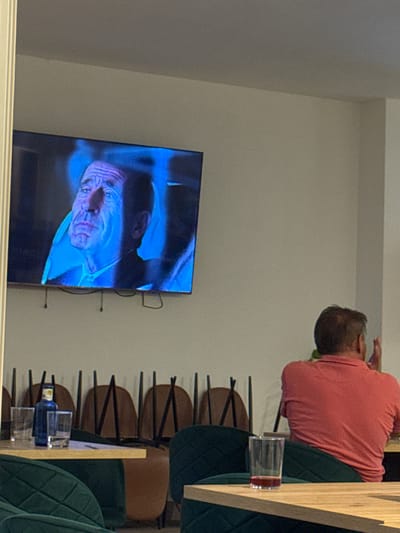 The image captures a scene from a restaurant in El Escorial, Spain, likely during the evening or night given the interior lighting. The main subjects are a person seated at a table and a television screen displaying a dramatic close-up of a person's face with intense blue lighting.
The seated person is viewed from behind, wearing a salmon-colored shirt. They appear to be looking towards the television, possibly engaged in watching the program or movie. Two glasses of liquid are visible on the table in front of them, one with a reddish tint and another with a clear liquid, suggesting they are dining or having drinks. The table itself is a light-colored wood, contrasting with the dark green upholstered chairs surrounding it. The chairs have a distinctive quilted pattern on their backrests.
The television screen is the focal point on the left side of the image. The image on the screen is a high-contrast, blue-toned close-up of an older person's face, with deep wrinkles and an open mouth, creating a somewhat unsettling or dramatic effect. The facial features are strongly lit, emphasizing the textures and contours of the skin.
The background shows more of the restaurant's seating area, with several more dark green chairs visible, arranged in rows. The overall ambiance appears to be that of a modern, well-lit dining establishment. There is no visible text in the image. The image is oriented vertically, suggesting it was taken with a phone held in portrait mode.