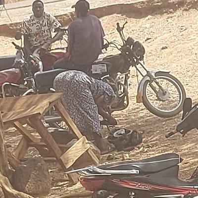 A woman in a floral dress sits on a motorcycle, facing away. To her left, a man in a purple shirt stands, while another man in a patterned shirt is seated on another motorcycle. A wooden structure, possibly a chair or stool, leans against the motorcycles. Additional motorcycles are parked nearby, and the background appears to be a textured, earthy surface.