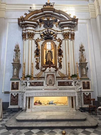 The image displays an ornate altarpiece and its surroundings within a church in Monopoli, Italy. The altarpiece, the central subject, is predominantly white with extensive gold leaf detailing, showcasing a Baroque or Rococo architectural style.

In the upper-middle section of the altarpiece, a framed religious painting or icon depicts a dark-robed figure, possibly a saint or monastic figure, standing within an interior. This central artwork is flanked and surmounted by elaborate gold and dark decorative elements, including scrolling volutes, carved angelic figures, and layered architectural forms.

Below the central painting, the altarpiece transitions to a marble base. To the right, a glass-fronted shrine or reliquary houses a recumbent sculpted figure, possibly Christ, made of a light-colored material and draped in blue fabric, resting on a red cloth. A delicate white lace curtain partially covers the opening of this shrine. Tall, decorative candelabras with multiple candleholders are positioned on either side of the main altar, along with other smaller, intricate architectural decorations.

The wall behind the altarpiece is a light blue-grey, complementing the white and gold. The floor in the foreground features a distinct checkered pattern of black and white marble tiles. Visible to the right are a simple wooden chair and a microphone stand, suggesting the space is actively used for services or events. The overall impression is one of grandeur and reverence, typical of historic church interiors. No discernible text is visible.