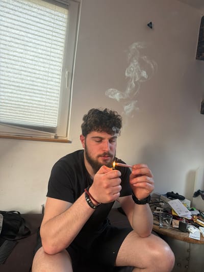 A man with curly hair and a beard is sitting indoors and lighting a rolled cigarette with a lighter. He is wearing a black t-shirt, shorts, a watch, and bracelets. Smoke is visible in the air. A window with blinds is in the background, and a table cluttered with various items is also visible. There is no indication in the image that the location is Lahnau, Germany.