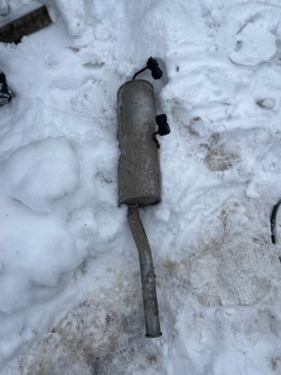 A detached automotive exhaust muffler with an integrated pipe section is depicted lying on snow-covered ground in Карабоўскі сельскі Савет, Belarus. The cylindrical muffler body, appearing metallic silver-grey, is covered in dirt and what appears to be corrosion. Two black electrical connectors, each secured by a short, bent metallic rod, are attached to the top surface of the muffler. The exhaust pipe extends from one end of the muffler, curving downwards, and is similarly soiled and corroded. The ground is covered by an uneven layer of white snow, interspersed with exposed patches of brown earth and mud, particularly around the object. A segment of a black cable or hose is partially visible on the far right edge of the frame. No individuals or active subjects are present.