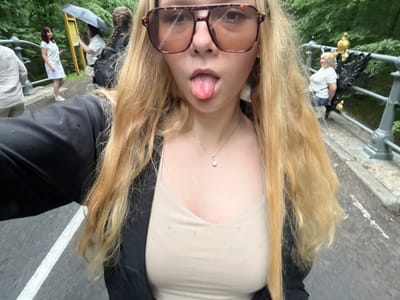 The image captures a close-up selfie of a young woman with blonde hair and large, light brown sunglasses, sticking her tongue out. She is wearing a light beige camisole and a dark jacket. A small, diamond-like pendant necklace is visible. In the background, several people are present on a bridge or walkway. To the left, a woman in a white dress is visible. Further back, another person holds an umbrella. To the right, a woman in a white t-shirt and jeans is seen standing near a decorative metal railing with golden and black accents. The environment appears to be outdoors, with greenery suggesting a park or forested area. The ground is paved, with a white line marking a path. The location is identified as Sharashėvski sel'ski Savet, Belarus.