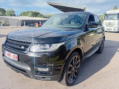 A black Range Rover Sport is parked on an asphalt surface, possibly a parking lot or rest stop, in front of a modern building with a unique angled roof. The building appears to be a service area or facility, with trees visible in the background. A white truck with "THU TRUCK" written on it is parked further back to the right. The Range Rover has a Belgian license plate displaying "2-ABS-428" and "CSC-BELGILIM.be". The sun is shining, casting shadows, indicating it is daytime. The vehicle is clean and appears to be in good condition, with a prominent "RANGE ROVER" badge on the hood and a Land Rover emblem on the grille. The overall scene suggests a brief stop during travel or a visit to the facility. There are no people clearly visible in the image.
