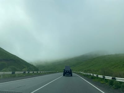 The image shows a rear view of a black RAM pickup truck driving on a paved road. The truck has a large, flat, dark object strapped to its bed, obscuring the view of the cab. The road is divided by a white line, and there is a guardrail on both sides of the road. The landscape consists of rolling green hills covered in grass, with fog obscuring the tops of the further hills. The sky is overcast and a muted gray-blue, suggesting a cloudy or foggy day. A second, smaller car is visible in the distance on the left side of the road. The overall mood is somewhat subdued due to the weather and the vast, empty landscape. There is some visible text on the license plate of the RAM truck, which appears to read "S363 BH 07".