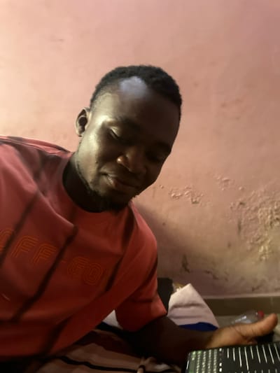 An adult male is depicted in an indoor setting in Agbogugu, Nigeria. He has dark skin, short dark hair, and a light beard. He wears a red short-sleeved t-shirt with "OFF-GO" branding. His gaze is directed downwards. His right hand holds a dark electronic device displaying a screen with vertical bars and symbols. He is situated on a striped fabric, with a white object and a partially visible clear plastic bottle to his right. The background is a light pink wall showing significant areas of peeling paint and discoloration, particularly in the mid-right section. A darker baseboard or floor is visible along the bottom edge of the wall.