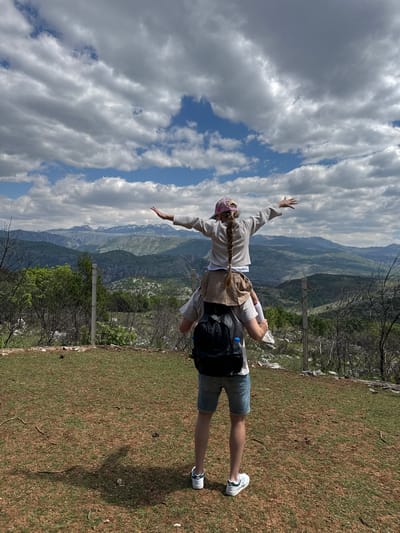 A man stands on a grassy and dirt-covered area, with a woman on his back appearing to be flying or mimicking an airplane. The location is Gornje Mrke, Montenegro, as indicated by the mountainous background and vegetation characteristic of the region. The man wears denim shorts and white sneakers, carrying a black backpack. The woman has a braid and wears a long-sleeved shirt, a light-colored skirt, and a pink hat or headband, with her arms extended outwards. In the background are mountains and a partly cloudy sky. A fence line and some sparse trees are visible.