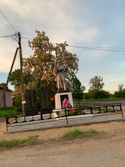 The image depicts a war memorial located near the village of Blevchitsy, within the Blevchitsy rural council of Belarus. The memorial features a statue of a soldier holding a flag, standing on a pedestal. Two dark stone tablets with inscriptions are situated in front of the pedestal, to the left. A wreath of colorful flowers is placed at the base of the pedestal. The memorial is enclosed by a black metal fence. Behind the memorial, a large tree with white blossoms is prominent. A utility pole with numerous wires is visible to the left of the memorial, and a pink building is partially visible in the far left background. The scene is set against a backdrop of trees, a road, and a sky with scattered clouds, suggesting an outdoor, rural setting during either sunrise or sunset due to the warm lighting.