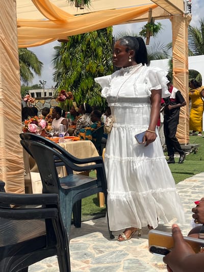 The image portrays an outdoor social gathering in Kumasi, Ghana. In the center-right foreground, a dark-skinned woman stands on a variegated stone-paved surface. She is wearing a white, floor-length, tiered, semi-sheer dress with ruffled short sleeves, a high collar, and horizontal dark gray bands on the bodice. Her dark hair is styled in a high ponytail with laid edges, and she wears pearl drop earrings. A thin necklace is visible, and she holds a dark-cased smartphone in her right hand. Gold-colored, embellished high-heeled sandals are on her feet, and a small patterned shoulder bag with a gold chain strap is visible on her left hip. She is facing slightly left, with her head turned to the left.
To the left, several dark plastic chairs are arranged near a round table covered with an orange tablecloth. Two floral centerpieces, containing pink, orange, and maroon flowers, are on the table alongside glassware and a water bottle. Multiple individuals are seated around this table, partially visible, some dressed in patterned attire.
In the mid-ground right, a man in a black long-sleeved shirt with a red and white shoulder panel and black trousers stands on artificial green turf. A woman in a yellow garment is partially visible behind him. Overhead, orange drapery fabric is stretched across a support structure, from which green leafy decorations are suspended. Lush green trees and shrubs form the background, with a stone wall visible behind the seated guests. An overcast sky is present above the foliage. In the lower right foreground, a hand holding a light-colored smartphone with visible camera lenses is partially depicted.