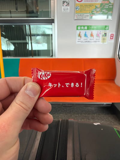 A hand holds a red, Japanese Kit Kat wrapper in an indoor setting, likely a train. The wrapper features the Kit Kat logo and Japanese text. The train interior shows an orange seat back, a window with a blurry reflection, priority seating signs, and luggage. The overall scene suggests a traveler enjoying a Kit Kat on a Japanese train.
