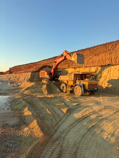 In an outdoor mining or construction setting, likely in Tatsinskaya, Russia, a large orange Hitachi excavator is actively digging and loading earth into a yellow and blue dump truck. The excavator's bucket is full, and it's in the process of dumping its load. The dump truck is positioned to receive the material, with its bed open. The scene is set against a clear blue sky, suggesting fair weather. The warm, golden light illuminates the excavated earth, casting long shadows and indicating that it is either early morning or late afternoon, with the sun low on the horizon. The ground is heavily textured with tire tracks from heavy machinery, signifying ongoing activity. The excavated earth shows distinct layers of different soil colors, ranging from reddish-brown at the top to lighter sandy tones lower down. The overall impression is one of active, large-scale earthmoving operations. Visible text on the excavator reads "HITACHI".