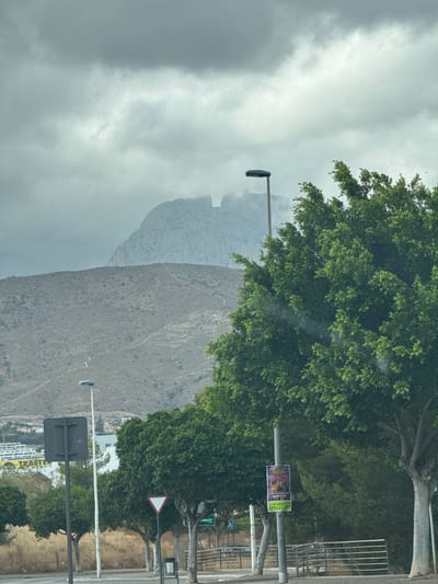 This media file depicts a view of a mountainous landscape, likely taken from a vehicle. The foreground is dominated by a large, lush green tree, its branches partially obscuring the view. To the right of the tree, there is a road with a sidewalk, marked by a white line and bordered by dry, yellow grass. Various street signs are visible, including a triangular warning sign and a rectangular sign with green text, though the text is mostly unreadable. A lamppost stands prominently in the foreground.
In the background, a vast, rocky mountain range rises dramatically. The mountain slopes are covered in sparse vegetation and crisscrossed by what appear to be walking paths. The upper portions of the mountains are shrouded in a thick layer of gray clouds, creating a somewhat dramatic and overcast atmosphere. The lighting suggests it is daytime, but the heavy cloud cover indicates overcast weather.
A small, colorful poster is attached to a lamppost, displaying the word "KPOP" prominently. Further in the distance, buildings can be seen clustered at the base of the mountains. The overall scene conveys a sense of being on the outskirts of a city, with nature and infrastructure coexisting. There are no people visible in the image. The orientation of the image is such that the landscape appears to be sideways, as if the camera was tilted.