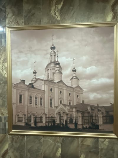 The sepia-toned photograph in a gold frame depicts a large, ornate church building with multiple domes and spires. The architecture suggests an Orthodox Christian style. The building is light-colored, possibly white or pale pink, with dark-trimmed windows. A decorative fence runs in front of the building. The sky behind is cloudy. The image is framed by marbled tile walls. This building is identified as the Holy Trinity Cathedral, located in Kursk, Russia.