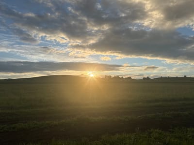 This is a landscape photograph captured at dusk or dawn, with the sun low on the horizon. The scene depicts a rolling, grassy field stretching towards a gentle, tree-lined hill in the distance. The sky is a dramatic mix of blue and grey clouds, illuminated by the bright, radiant sun. The sun's rays are casting a warm, golden glow across the entire scene, creating a hazy, ethereal effect. The foreground features a dark, tilled patch of earth with some green grass growing at the very bottom. The overall mood is serene and peaceful, evoking a sense of natural beauty and tranquility. The location is identified as Elimbetovo, Russia. There is no visible text in the image.