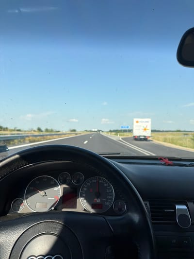 The image captures a first-person perspective from inside a vehicle, likely an Audi RS6 given the visible logo on the speedometer, traveling on a multi-lane highway under a clear blue sky. The scene is set in the vicinity of Svoboda, Bulgaria, during daylight hours.
In the immediate foreground, the top portion of a black, leather-wrapped steering wheel with an Audi logo is visible. Behind the steering wheel, the instrument cluster displays a tachometer on the left indicating approximately 2000-2500 RPM and a speedometer on the right showing a speed of around 100-110 km/h. Other gauges for fuel, engine temperature, and possibly oil are also present. To the right of the steering wheel, a part of the dashboard is visible, featuring an air vent with an attached silver magnetic phone mount. Below the vent, buttons labeled "ESP" and "P" are discernible. A small, red, possibly reptilian-shaped ornament rests on the dashboard near the base of the windshield on the right side. A rearview mirror is partially visible in the top right corner. The windshield itself is marked with numerous small streaks and spots, indicative of dirt, dust, or insect residue.
Beyond the windshield, a straight, asphalt highway stretches into the distance under a vast, mostly cloudless blue sky, with a few faint wisps of clouds on the horizon. The highway has white solid lines marking the shoulder and lane divisions. Green grassy verges line both sides of the road, with some trees and bushes further back, suggesting a rural or semi-rural environment. Ahead and slightly to the right, a white box truck with a colorful logo featuring a star-like design and text that appears to be "HOLiME" is visible on what looks like an exit ramp or merging lane. To the left of the truck, a blurred blue rectangular road sign is present. No other prominent vehicles are in the immediate vicinity.