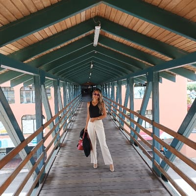 A woman stands centered on a covered wooden bridge, wearing a black tank top and white pants. She holds a red bag and a black coat on her left arm. The bridge's structure features teal-colored beams and railings, with a wooden roof and exposed wood planks forming the walkway. Buildings with windows are visible on either side of the bridge, and the bridge stretches into the distance. An overhead light hangs from the roof.