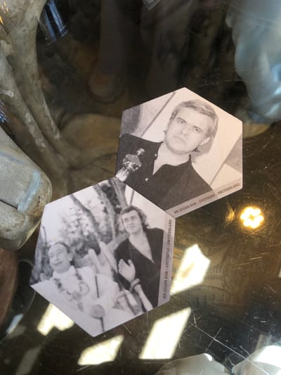 The image shows two hexagonal coasters featuring black and white images and text that reads "HR GIGER BAR - GRUYERES - SWITZERLAND." One coaster displays a portrait of a man with a serious expression, holding what appears to be a sculpted object. The other coaster shows a blurry image of several people at a table, potentially in a bar setting. The coasters are placed on a reflective surface which appears to be the top of a glass-encased display or table. The reflection shows light sources and objects within the display case. The overall scene appears to be inside the HR Giger Bar in Gruyères, Switzerland, a location known for its surreal and biomechanical design inspired by the artist H.R. Giger.