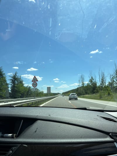 A view from inside a car driving on a highway. The driver is traveling on the right lane. A white car is visible ahead in the same lane. A speed limit sign indicating 100 is visible on the right side of the road, along with a hazard warning sign and a snow warning sign. The sky is blue with scattered clouds. The location is along a highway, and there is no evidence that this is Slavovitsa, Bulgaria. The interior of the car, including the dashboard and vent, is visible on the right. A lighter is visible in a compartment within the dashboard. The windshield has some smudges.