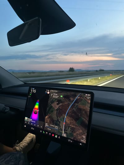 The image depicts the interior perspective from the driver's seat of a Tesla vehicle traveling on the A4 Maritsa highway, near Georgi-Dobrevo, Bulgaria, during sunset or dusk. The sky above is light blue, transitioning to hues of orange and pink near the horizon, with distant hills visible. The multi-lane highway stretches forward, illuminated by orange reflector markers on the right shoulder.
Inside the car, a central vertical touchscreen displays vehicle and navigation information. The left panel shows the car traveling at 115 km/h, with a green headlight indicator, and a graphical representation of the car on a rainbow-colored road. Below this, a music player interface is active, showing "MLADO DAZE" by "MOMO RECORDS, Krisko" playing, with a volume level of "21.5". The right panel displays a map indicating "50 km" to Dimitrovgrad, Bulgaria. Vehicle status information includes "72%" battery charge, time "21:12", and an ambient temperature of "26°C". Navigation details show "2 hr 40 min" and "189 km" remaining to "End Trip" at Kilifarevo, Bulgaria, with the current mapped location indicating "A4 Любимец" (Lyubimets). A Shell station icon is also visible on the map.
In the bottom left, a person's left foot, wearing a light-colored sneaker, rests on the floor mat, with part of a patterned garment visible on the leg. A dark dashboard extends across the foreground. A rearview mirror is partially visible at the top, reflecting the sky.
