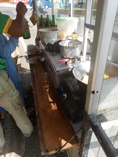 The image depicts an outdoor scene at a street food stall in Ruwan Eliya, Sri Lanka. A person, partially visible from the waist down, is standing near the stall, holding a green glass bottle. They are wearing khaki pants with multiple pockets and dark footwear. The stall itself appears to be constructed of metal and wood, with various food preparation items visible. These include pots and pans, one of which contains yellow food items, a portable stove, and containers for ingredients. A wooden plank extends from the stall, holding a glass with a brown liquid and what appears to be a mortar and pestle on a small plate. In the background, a blue vehicle is parked on a street, and other elements of an urban environment are suggested, such as buildings and foliage. The overall impression is one of a busy street-side food vendor preparing or serving.