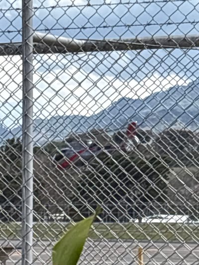 The scene presents an outdoor view through a chain-link fence, which occupies the immediate foreground, featuring a grey vertical support post on the left and a horizontal top rail. A green leaf is visible at the bottom left. In the mid-ground, a helicopter, predominantly dark-colored with prominent red markings on its tail section, rests on the ground within an open paved or grassy area. Beyond the helicopter, a line of trees precedes a series of distant, undulating mountains under a partly cloudy sky. No individuals are discernible. This setting is consistent with the perimeter of Tobías Bolaños International Airport in Pavas, San José, Costa Rica, facing the surrounding mountainous terrain of the Central Valley.