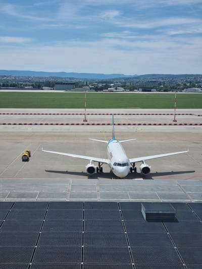 An aerial view captures an aircraft parked at Stuttgart Airport, located in Echterdingen, Germany. The plane, predominantly white with blue and red accents, rests on a tarmac marked with red and white striped barriers. Adjacent to the plane are rows of solar panels. In the background lies a cityscape, with clear blue sky.