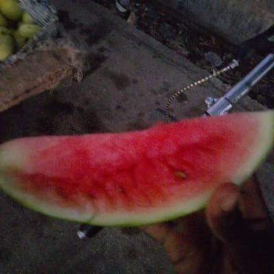 The image shows a close-up of a slice of watermelon. The watermelon is bright red with some visible seeds and a pale green rind. A person's hand is partially visible on the right side of the image, holding the watermelon. In the background, there is a rough, dark surface, possibly concrete or asphalt, with some debris scattered on it. To the left of the watermelon, there appears to be a brown, textured material, possibly burlap or a woven bag, and some round, yellow objects that could be other fruits or vegetables. A metal object, possibly a part of a tool or equipment, is also visible in the upper right portion of the background, with a thin, coiled wire extending from it. The overall lighting suggests it could be daytime, but the quality of the image makes it difficult to determine precise details about the environment or the time of day. The setting appears to be an outdoor, possibly a market or street stall, given the presence of produce and the rough ground. There is no visible text in the image.