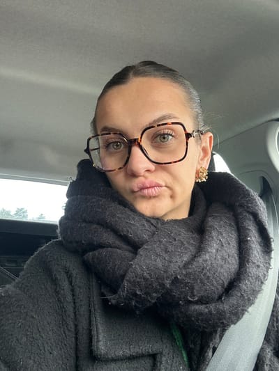 A woman is depicted seated inside a vehicle in Saint-Cyprien, France, posing for a photograph. She has light eyes, light skin, and her dark hair is pulled back. She wears large, tortoise-shell framed eyeglasses, a gold-toned floral stud earring on her left ear, a thick dark-colored coat, and a voluminous dark scarf wrapped around her neck. Her lips are pursed in a pout, and she looks directly towards the camera. A light-colored seatbelt crosses diagonally across her chest. The vehicle's gray fabric headliner is visible above her, and through the window behind her, an indistinct outdoor scene of an overcast sky and distant foliage is partially visible.