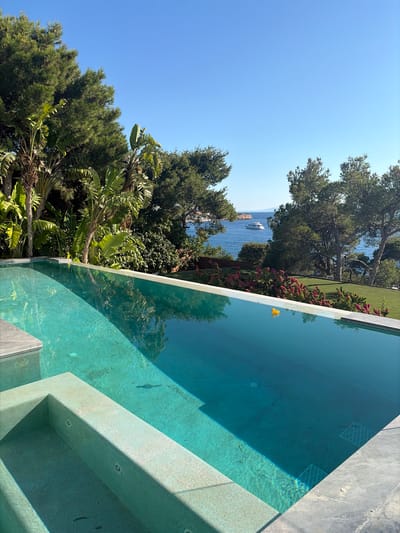 An infinity swimming pool with clear turquoise water dominates the foreground of the image. Steps are visible within the pool's shallow end. The pool is surrounded by a stone coping. Beyond the pool, lush green trees and tropical plants create a dense canopy. In the background, the azure blue sea stretches out to the horizon, dotted with a white yacht. A distant island is visible under a clear blue sky. The location is the city of Vouliagmeni Municipal Unit, Greece. There are no people in the image.