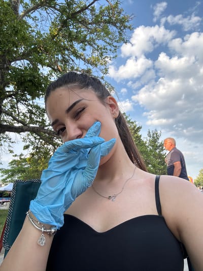 An outdoor close-up shot captured in the city of Kostievo, Bulgaria, features a young female subject prominently in the foreground. She has dark hair styled back and is winking with her left eye. Her right hand, covered by a light blue disposable glove, is placed against her cheek, partially obscuring her mouth. She is wearing a black strapless top or dress, a silver necklace with two small charms, and a silver charm bracelet on her left wrist. A segment of a green fabric chair is visible in the lower left. The background consists of a clear blue sky with scattered white cumulus clouds and abundant green tree foliage, including a large deciduous tree to the left. In the mid-ground on the right, an older male with grey hair, wearing a dark blue polo shirt, is partially visible from the chest up, facing away from the camera. A white tent or canopy structure is faintly discernible in the far left background.
