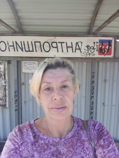 The image shows a woman with blonde hair, wearing a purple shirt and a necklace. She is standing in front of a sign that reads "КОНШПОРТНА" in large, dark letters, with some graffiti over it. To the right of the sign is a blue and red emblem. Behind the woman, there is a corrugated metal wall with some writing on it. The scene appears to be an outdoor public space, possibly a bus stop or a train station, given the sign and the shelter-like structure above. The woman is taking a selfie, as her face is the primary focus of the image. The overall impression is casual and informal. Based on the text on the sign, the location is likely the city of Kommunar, Russia, which is known for its industrial history and its administrative center, the settlement of Kommunar.