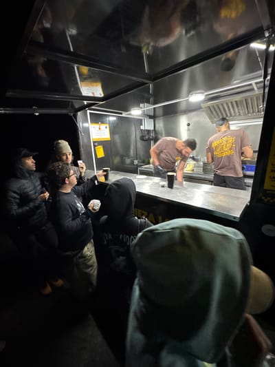 A view looking into a food truck. Two workers stand inside the truck, preparing food at a counter. Outside, three people are lined up waiting for their order, holding food and beverages. The location is San Bartolomé, Spain.
