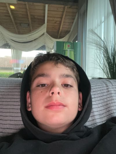 A close-up portrait shows a young person, likely male, reclining indoors and looking directly at the camera. The subject wears a black hooded sweatshirt with the hood up, framing their face. They have short, brown hair and dark brown eyes, with a neutral to slightly contemplative expression. The person appears to be resting on a grey fabric with subtle stripes, possibly a sofa or cushion.
The background is dominated by reflections in a window behind the subject. These reflections reveal elements of both an interior and exterior scene. A prominent feature is a green sign with a red star and the inverted, partially visible text "ekeneH," which is the Heineken logo, suggesting a commercial establishment such as a bar, pub, or restaurant. Other reflections include wooden paneling or ceiling structures, white drapes or fabric elements, and a plant with long, thin leaves. In the distance, reflections show tiled roofs and faint outdoor structures, indicating a broader setting. A subtle rainbow-like glare is also visible in the reflections.
The bright reflections suggest the photograph was taken during daytime. The scene depicts a casual indoor or covered outdoor area, likely within a commercial venue in Cherni Vrah, Bulgaria. The activity is a simple pose for a photograph, capturing a moment of repose.