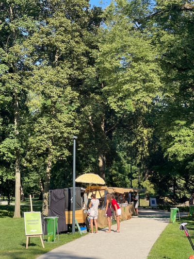 The image captures an outdoor market stall setup in a park-like setting, with lush green trees and manicured lawns suggesting a sunny day. The stall is positioned on a paved pathway, likely in Stupava, Slovakia, as indicated by the signage.
Three individuals are visible near the stall. Two younger women, dressed in casual summer attire, appear to be working at the stall. One is wearing a red top and white shorts, while the other is in a dark purple skirt and black top. A third person, a man in white shorts and a light-colored shirt, is also present, possibly a vendor or customer.
The stall itself is covered by a light-colored awning, and beneath it, a collection of items are displayed on tables, possibly food or artisanal products. There's a large yellow, straw-like umbrella leaning against the stall. Several black tarpaulins are draped over equipment, and a bright green trash receptacle is situated nearby.
A prominent sign on an easel in the foreground displays green text on a white background. It reads "REGIONÁLNE ŠPECIALITY" (Regional Specialties) with a logo that appears to be a green leaf or sprout. The overall atmosphere suggests a pleasant outdoor event, likely a local market or fair, taking place on a bright, clear day.