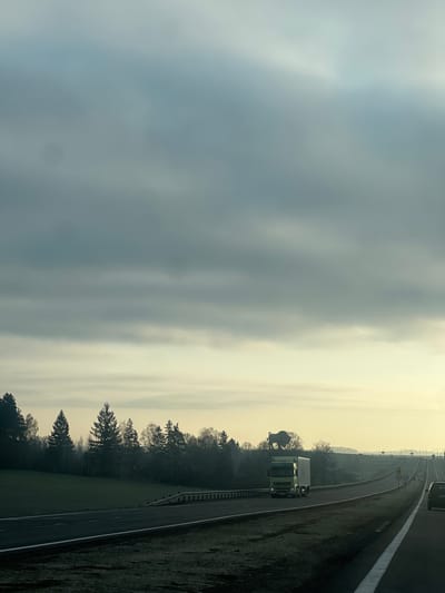 The image depicts a highway on the outskirts of Крошынскі сельскі Савет, Belarus. A truck travels on the road. The sky is overcast with a hazy, diffused light. Trees line the edge of the highway. A speed limit sign is visible in the distance.