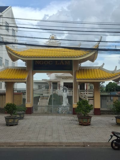 This is a medium-shot, eye-level photograph of the entrance gate of a Buddhist temple in B'Lao, Vietnam. The gate is adorned with a vibrant yellow, tiered roof, characteristic of traditional Vietnamese architecture. Above the gate, a circular Dharma wheel symbol is visible, representing Buddhism. The words "NGỌC LÂM" are inscribed in large, dark letters on a sign mounted on the gate, identifying the temple. Below this, smaller text reads "639 TRAN PHU THI X. BAO LOC." Two potted trees with green foliage are placed on either side of the gate, on a tiled walkway. Behind the gate, a metal fence encloses the temple grounds, with glimpses of buildings and more greenery visible. The scene is framed by power lines running across the top of the image and a cloudy sky. In the foreground, a portion of a road and a motorcycle are visible.