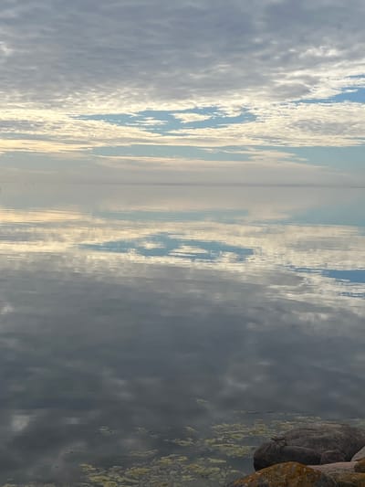 The image shows a tranquil scene at the coastline of Nasva alevik, Estonia. The sky is filled with scattered clouds in varying shades of gray and white, with hints of blue peeking through. The water's surface is calm and reflective, mirroring the sky and clouds, creating a sense of depth and expanse. In the foreground, rocks are visible along the water's edge, some partially submerged. A small amount of light-colored debris or vegetation is present on the water's surface and near the rocks. The overall atmosphere is peaceful and serene, suggesting early morning or late evening light. There are no people or animals visible in the image.