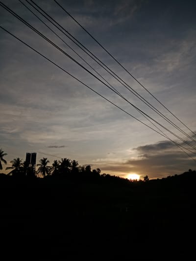 The image depicts a sunset scene in Seuneubok, Indonesia. Overhead, a series of parallel power lines stretch diagonally across the frame against a sky filled with striated clouds. The sun is visible near the horizon, obscured by a dark band of clouds. Below, the silhouetted forms of trees, including palm trees, create a dark foreground. Two vertical structures resembling silos are also visible. The overall scene is dark, emphasizing the sunset and the silhouetted landscape.