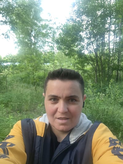 A mid-shot, front-facing selfie captures a person with dark, short hair and fair skin, wearing a yellow and dark blue jacket with a grey hood. The person is outdoors, with a blurred background of green trees and foliage, indicative of a natural environment. The setting appears to be a park or wooded area, potentially in or near the village of Tsibino, Russia. The person is looking directly at the camera with a neutral expression, and a black earbud is visible in their right ear.