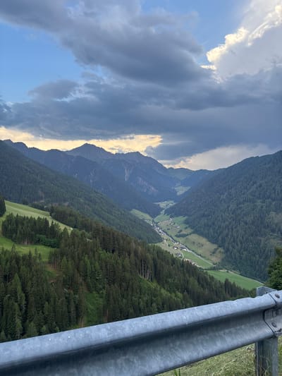 This is a vertical, eye-level, medium shot of a mountain valley in Ratschings - Racines, Italy. The sky is dramatic with dark, heavy clouds on the left, interspersed with bright, sunlit patches of sky on the right. The light suggests either sunrise or sunset.

The valley floor is visible, with a small village nestled among green fields and dark, dense pine forests that climb the steep slopes of the mountains. The mountains themselves are layered and extend into the distance, with the furthest peaks appearing hazy. A metal guardrail runs along the right side of the frame, suggesting the vantage point is from a road or walkway overlooking the valley. The overall impression is one of natural beauty and a sense of peaceful isolation, with the dramatic sky adding an element of awe. There is no visible text in the image.