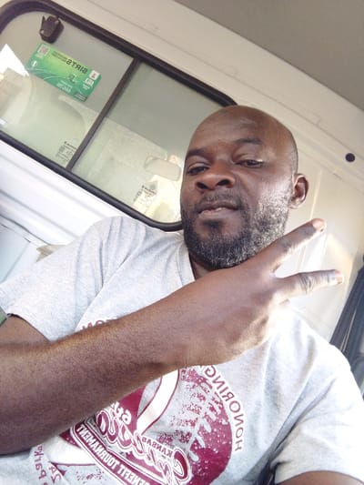 A bald, Black man with a beard and mustache is seated, wearing a grey t-shirt with red and white graphics. He is making a "W" or "peace sign" with his hand. He appears to be inside a vehicle. The vehicle’s window is visible behind him, with a sticker and rearview mirror in view. The interior of the vehicle appears to be white.