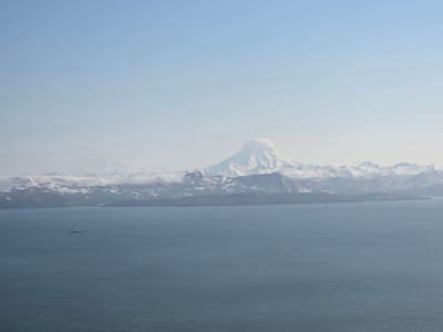 This is an aerial wide shot of a vast expanse of calm, dark blue ocean in the foreground. In the mid-ground, a body of land is visible, characterized by rugged, snow-capped mountains that stretch across the horizon. A plume of white smoke or cloud emanates from the peak of a prominent, conical volcano in the center of the mountain range. The sky above is a pale, clear blue with a gradient fading to white near the horizon. A small, dark ship with a white superstructure is positioned in the ocean to the left of the frame. The location is identified as being near the city of Krutoberegovy, Russia.