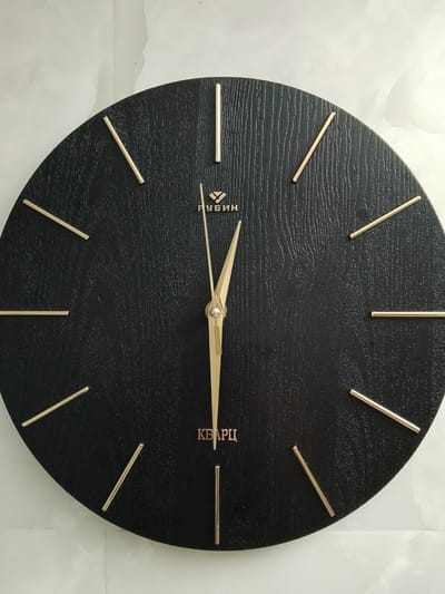 The image shows a close-up, eye-level view of a modern, minimalist black wall clock. The clock has a textured wooden face and features simple, gold-colored hour markers that are thin, straight lines. The hands of the clock are also gold-colored and appear to be made of metal. The hour hand is positioned slightly past the 12, and the minute hand is pointing towards the 2. The word "РУБИН" (Rubin) is visible in gold lettering above the center of the clock, and "КВАРЦ" (Quartz) is visible below the center. The clock is mounted on a light-colored wall with a subtle marbled pattern. There are no people or other objects in the image. The city of Yelo, Russia is not depicted or referenced in this image.