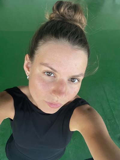 A young woman with her hair in a bun is looking up at the camera. She is wearing a black top and has several earrings in her left ear. The background is a solid green surface. The image appears to be a selfie taken outdoors. The location is not discernible from the image itself and is therefore not included.