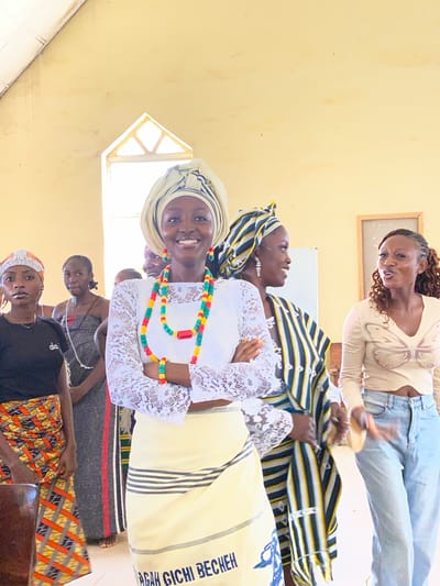 This media file shows a group of people in what appears to be an indoor gathering in Dutsen Kato, Nigeria. The setting is simple, with light-colored walls and a window with a wooden frame visible on the left. A framed picture hangs on the wall at the top. The people are dressed in a mix of traditional and contemporary clothing.
The primary focus is on a woman in the foreground, wearing a white lace top, a headwrap, and a colorful beaded necklace. She is smiling broadly and looking towards the camera. Behind her, other individuals are also visible, some wearing headwraps and patterned fabrics, suggesting a cultural event or a community meeting. One woman in the background is wearing a light-colored top and jeans, engaged in conversation or an activity. Another person is wearing a black t-shirt and holding a colorful piece of fabric.
The overall mood seems lively and engaging, with several individuals looking at the camera or interacting with each other. The natural lighting suggests it might be daytime. The presence of traditional attire and the location context point to a cultural gathering specific to Nigeria. There is some text visible on a yellow cloth draped over one of the individuals, which reads "RAH GICH BECH".
